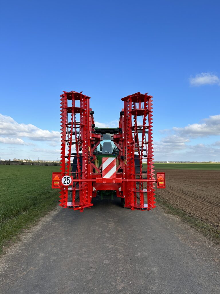 Vu arrière d'un cultivateur 3 poutres replié Gyrax. Attelé à l'arrière d'un tracteur sur une petite route.