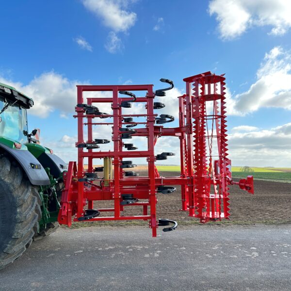 Vu côté d'un cultivateur 3 poutres replié Gyrax. Attelé à l'arrière d'un tracteur sur une petite route.