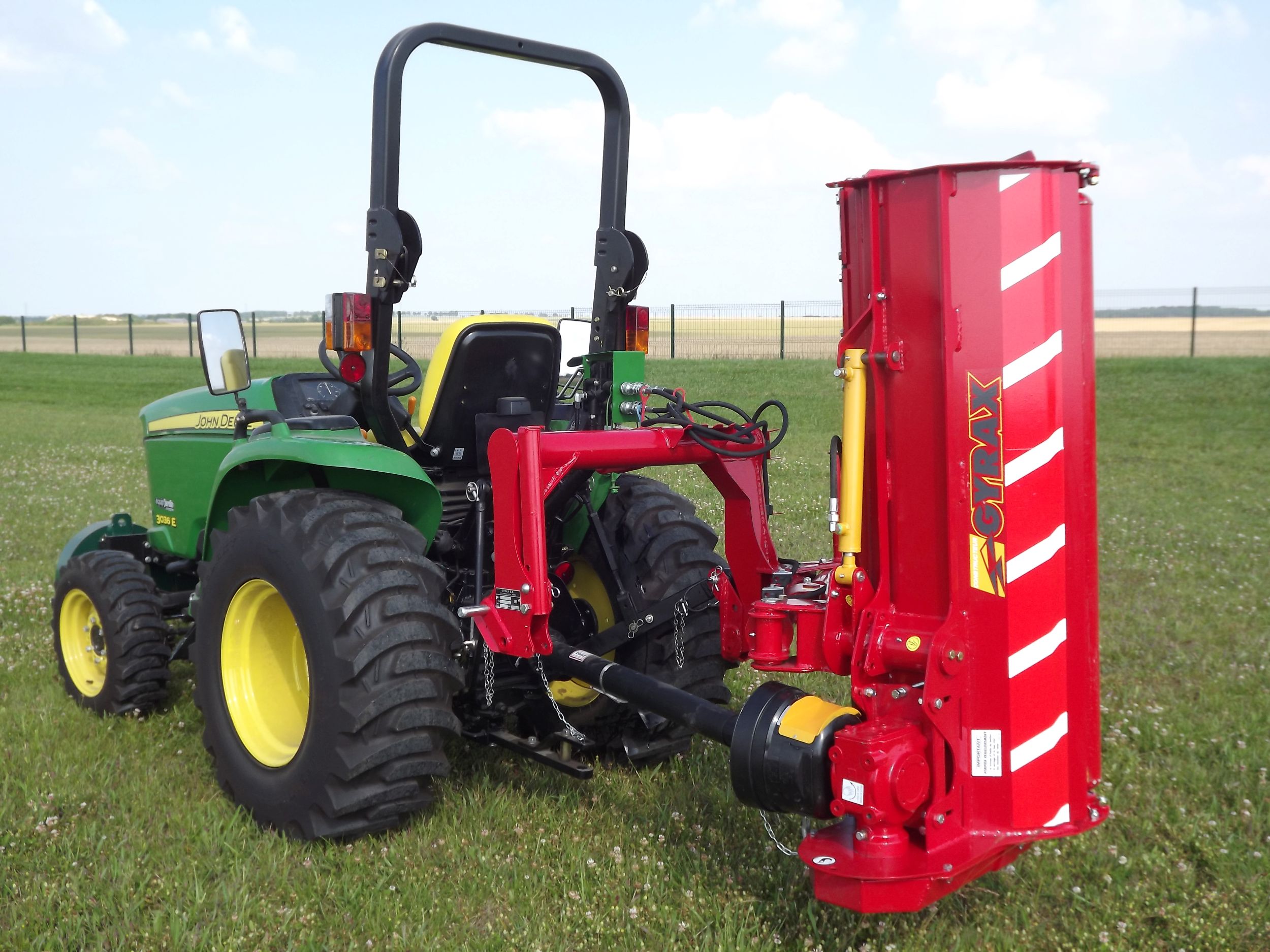 Roto-faucheuse rouge Gyrax, modèle RFL (loisir), vue 3/4 arrière attelée sur un micro tracteur, au transport dans un champs.