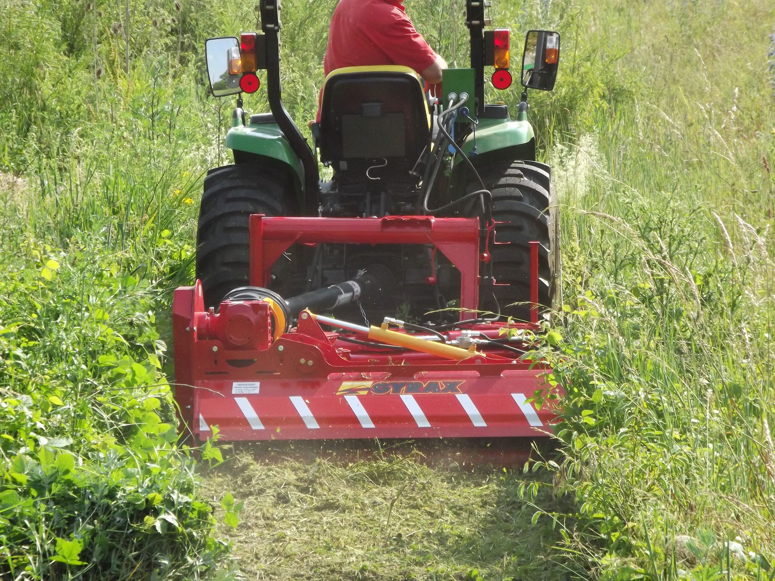 Roto-faucheuse rouge Gyrax, modèle RFL (loisir), vue 3/4 arrière attelée sur un micro tracteur, coupant des herbes tres hautes dans un champs. position repliée à l'arrière du tracteur en horizontal